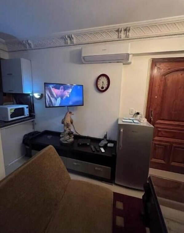 A compact living space with a television mounted on the wall, a microwave, a refrigerator, and a small table with various items.