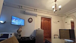 A living room with a television, a clock, a wooden door, and a desk with a computer. - Amazing studio apartment gleem (Alexandria)