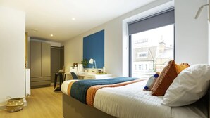 A modern bedroom with a large bed, a desk, and a window with a city view. - Student Only Market Quarter (Bristol)