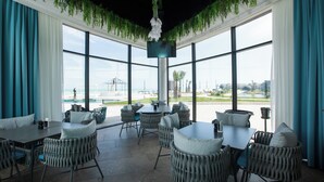 Amplio espacio interior con grandes ventanales, mobiliario de mimbre y vistas a la playa y al paisaje urbano.