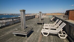 Terraza de madera con sillones y mesas, con vistas a una masa de agua y un puente distante.