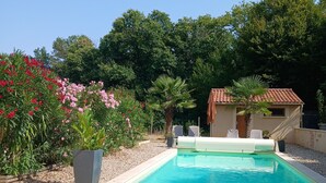 Ein Pool mit gefliestem Sonnendeck, umgeben von Kübelpflanzen und einem kleinen Gebäude mit einem Ziegeldach.