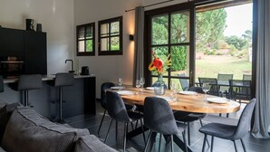 Un espace repas moderne avec une table en bois dressée pour quatre personnes, entourée de chaises noires. On aperçoit, à gauche, un coin cuisine avec un meuble sombre et une machine à café. De grandes fenêtres offrent une vue sur un jardin verdoyant à l’extérieur.