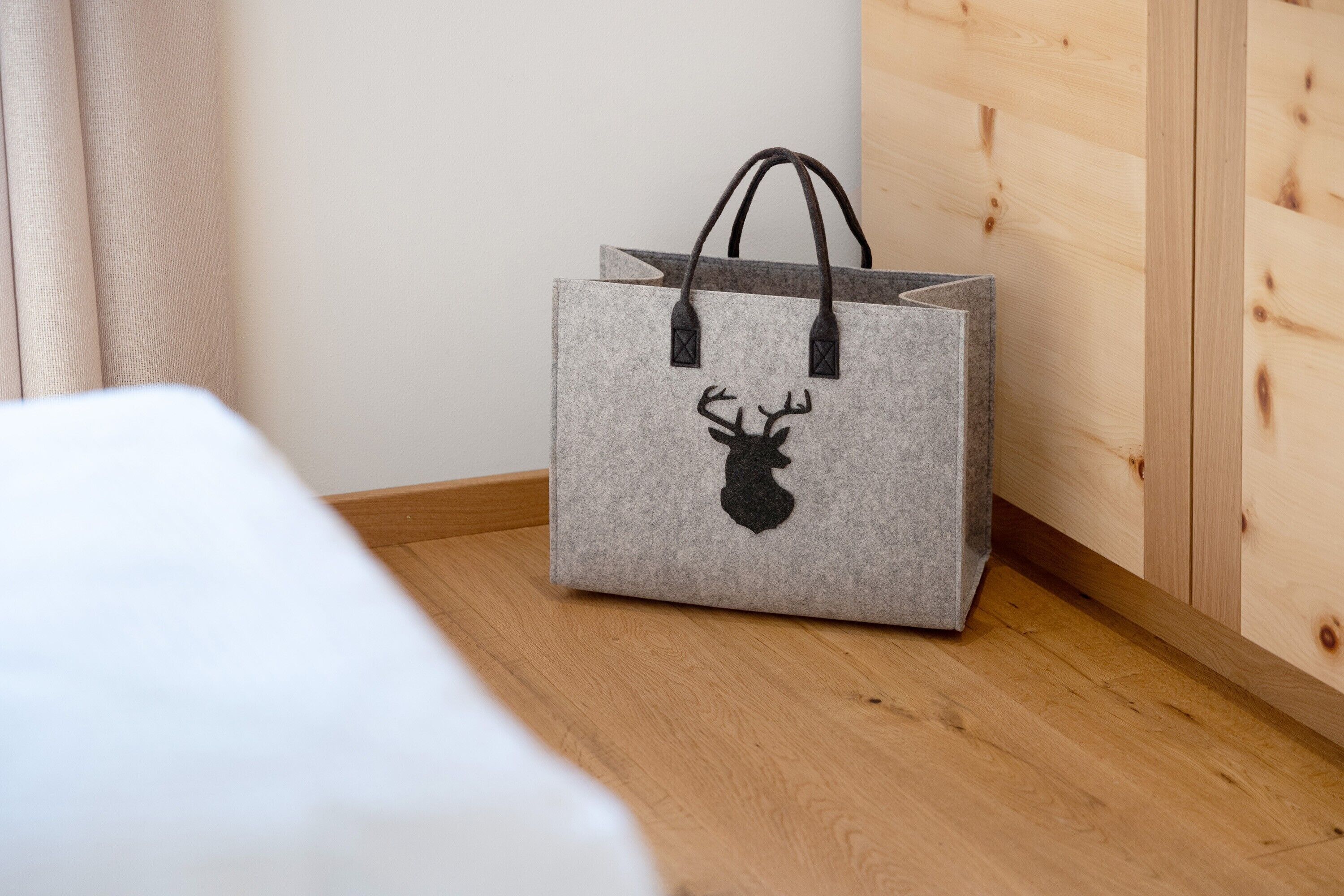 Una bolsa gris de fieltro con la silueta de un ciervo sobre un suelo de madera, junto a una cama blanca y una pared de madera.