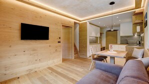 A modern interior with a wooden floor, a flat-screen TV mounted on the wall, a dining area with a table and chairs, and a kitchen in the background. - Apartment 'Weissbrunn Lisa' with Mountain View, Shared Terrace and Wi-Fi (St. Maddalena)