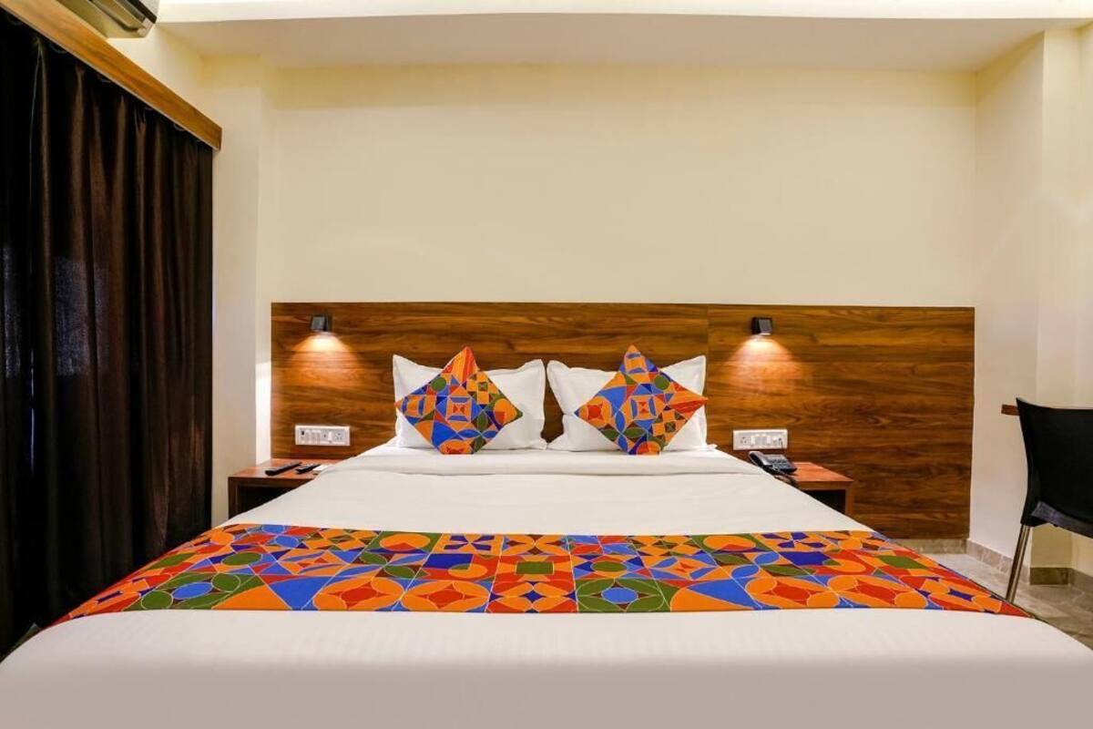 Quarto de hotel com cabeceira de madeira, duas luminárias de apoio, cama com colcha de padrão geométrico colorido e cortina escura à esquerda.