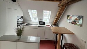 Una cocina compacta con armarios blancos, techo con vigas de madera, una pequeña mesa de comedor y piso rojo.