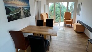 Un comedor moderno con una mesa y sillas de madera, un cuadro grande de un paisaje, y un balcón con vistas a zonas verdes.