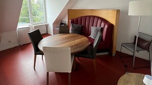 Una habitación con techo inclinado, una mesa redonda de madera con sillas, un cabecero tapizado en rojo y una lámpara de pie.
