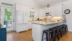 A modern kitchen with white cabinetry, a central island with bar stools, a clock on the wall, and a sign reading "COAST ALONG". - 4 BR Cottage w/ Pool, Hot Tub, Tiki Bar, 2 King Suites - Walk to Beach (Emerald Isle)
