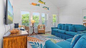 A living room with a blue sofa, a wooden coffee table, a wicker chair, and a television on the wall. - 4 BR Cottage w/ Pool, Hot Tub, Tiki Bar, 2 King Suites - Walk to Beach (Emerald Isle)