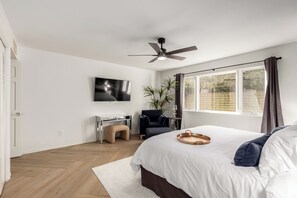 Un dormitorio con una cama, un escritorio, una silla, un ventilador de techo, un televisor y una ventana con cortinas.