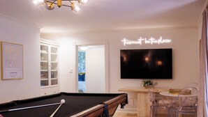 Et biljardbord i et rum med en lysekrone, et fladskærms-TV og et spiseområde med transparente stole.