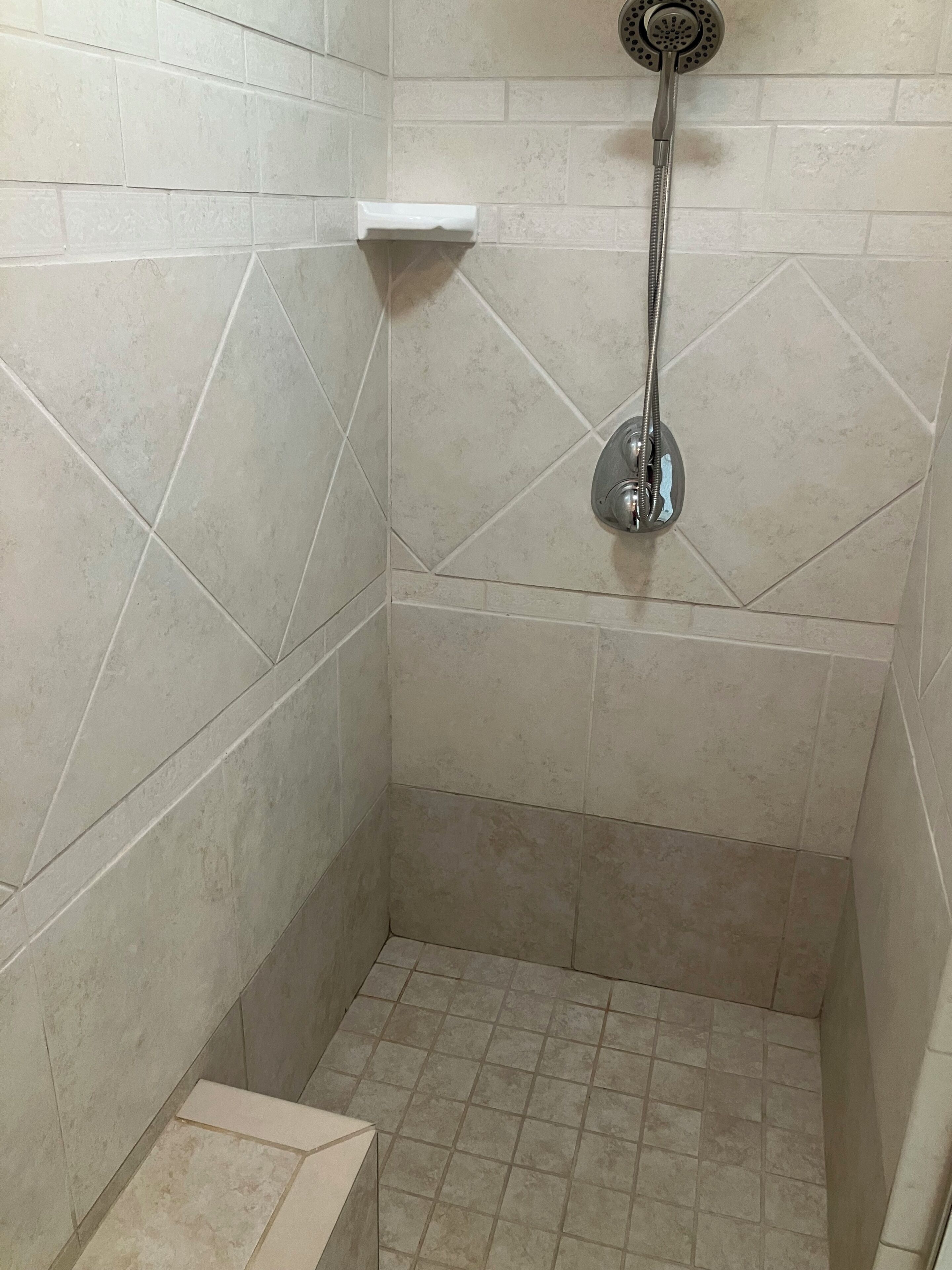 Ducha con cabezal de ducha de mano, estante blanco y paredes y suelo con baldosas.
