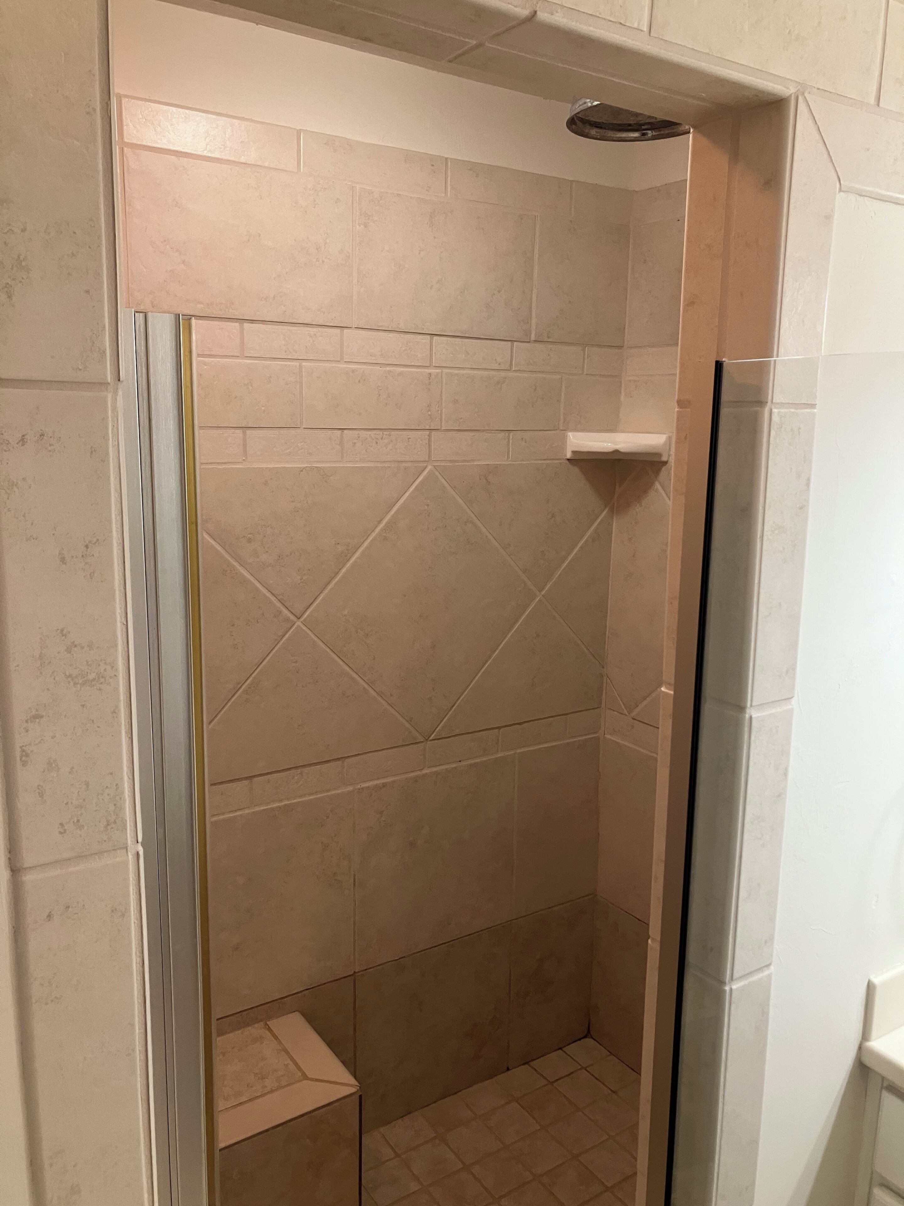 A shower with beige tiles, a glass door, and a white shelf.