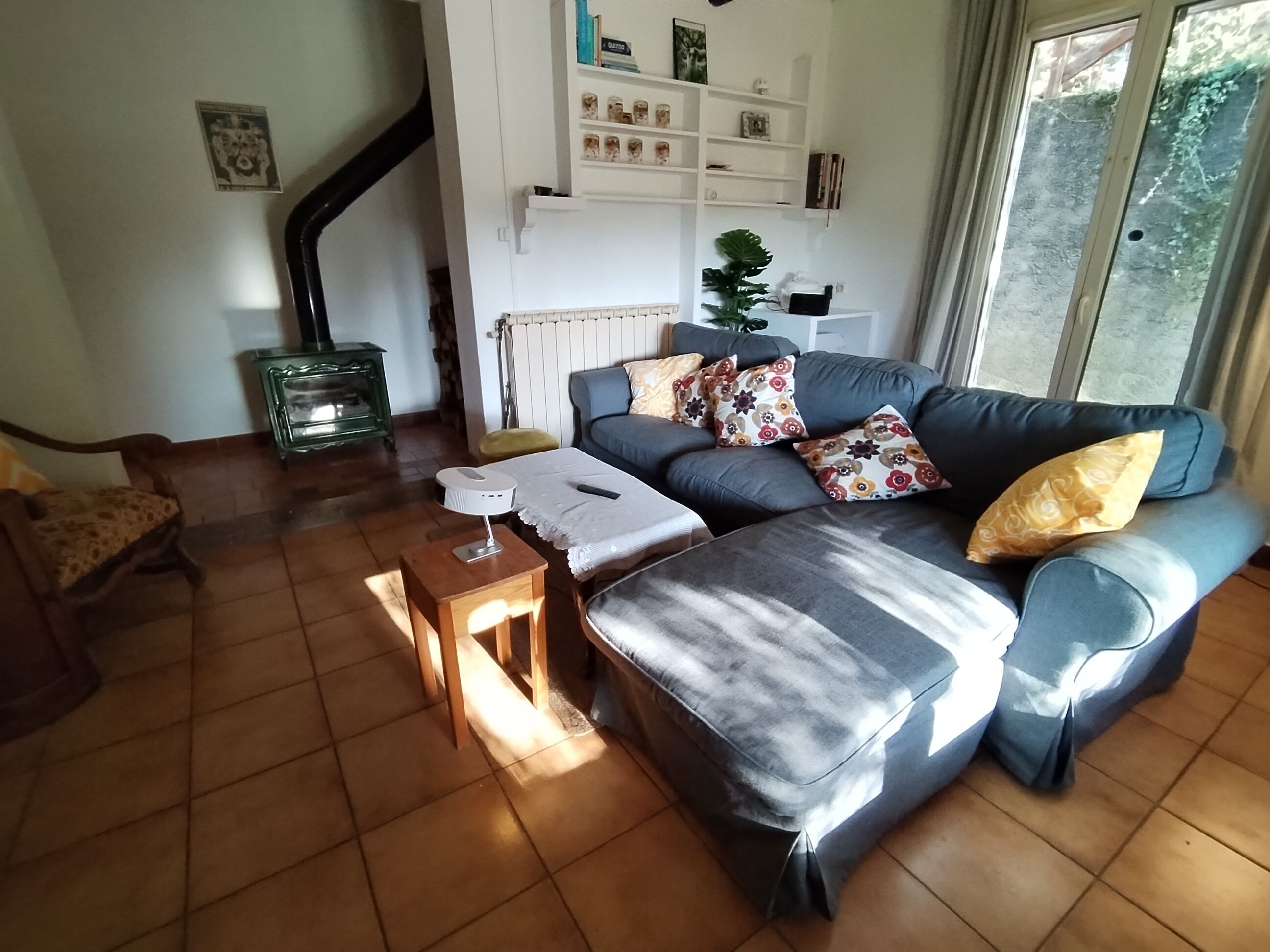 A living room with a sofa, a small table, and a radiator. There is a staircase in the background and a window with curtains.