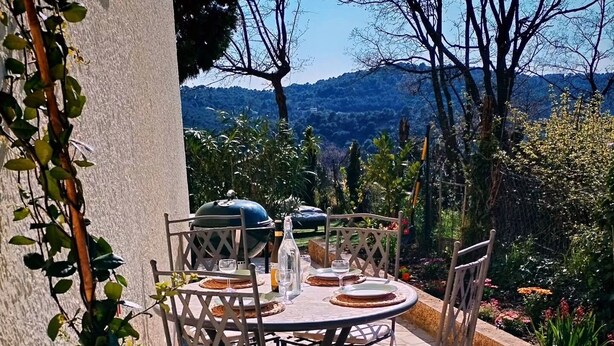 An outdoor patio with a round table and chairs, surrounded by potted plants and a view of hills.