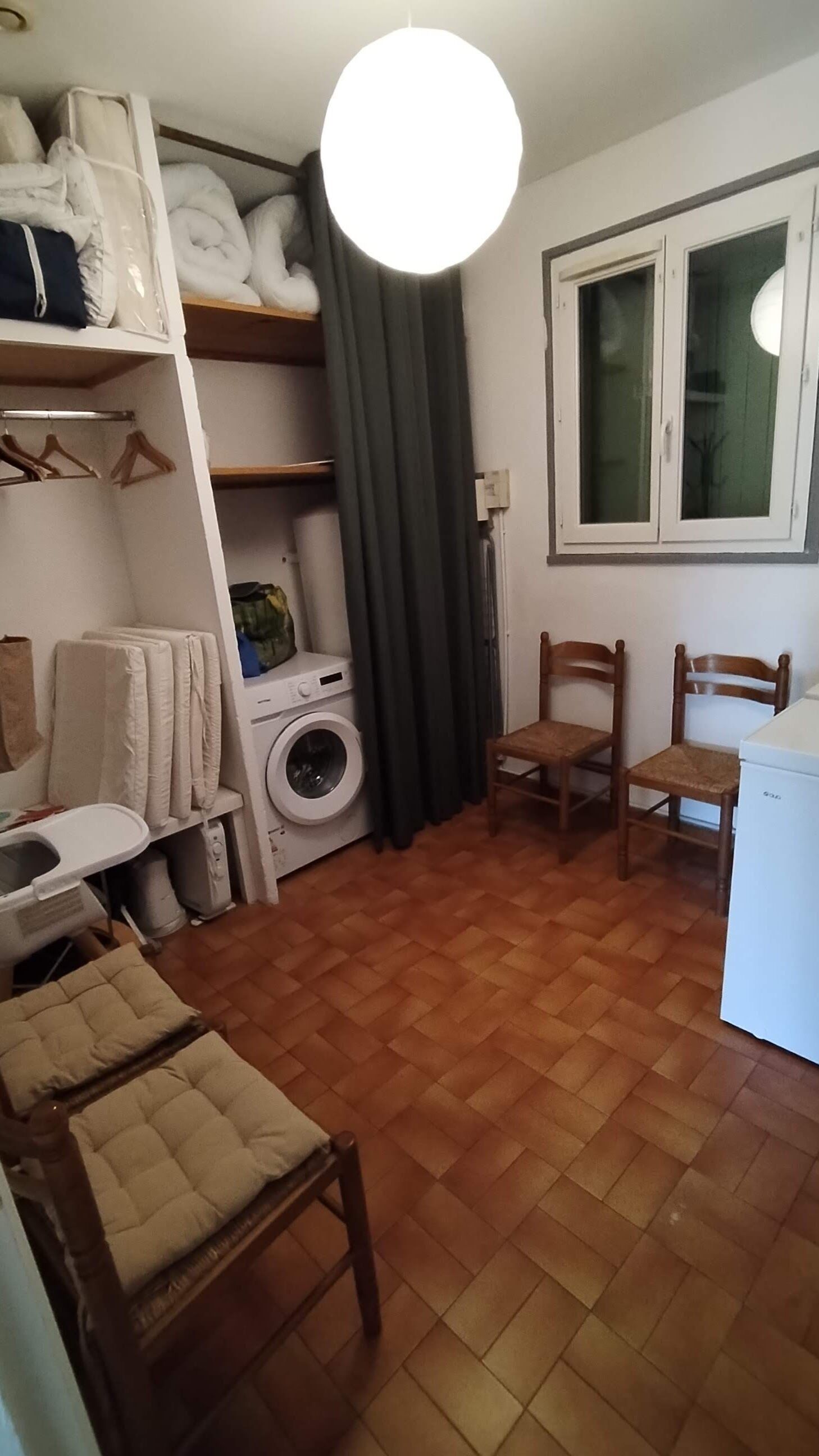 Un espace de vie compact comprenant un lave-linge, une chaise, une petite table et une fenêtre avec des rideaux.