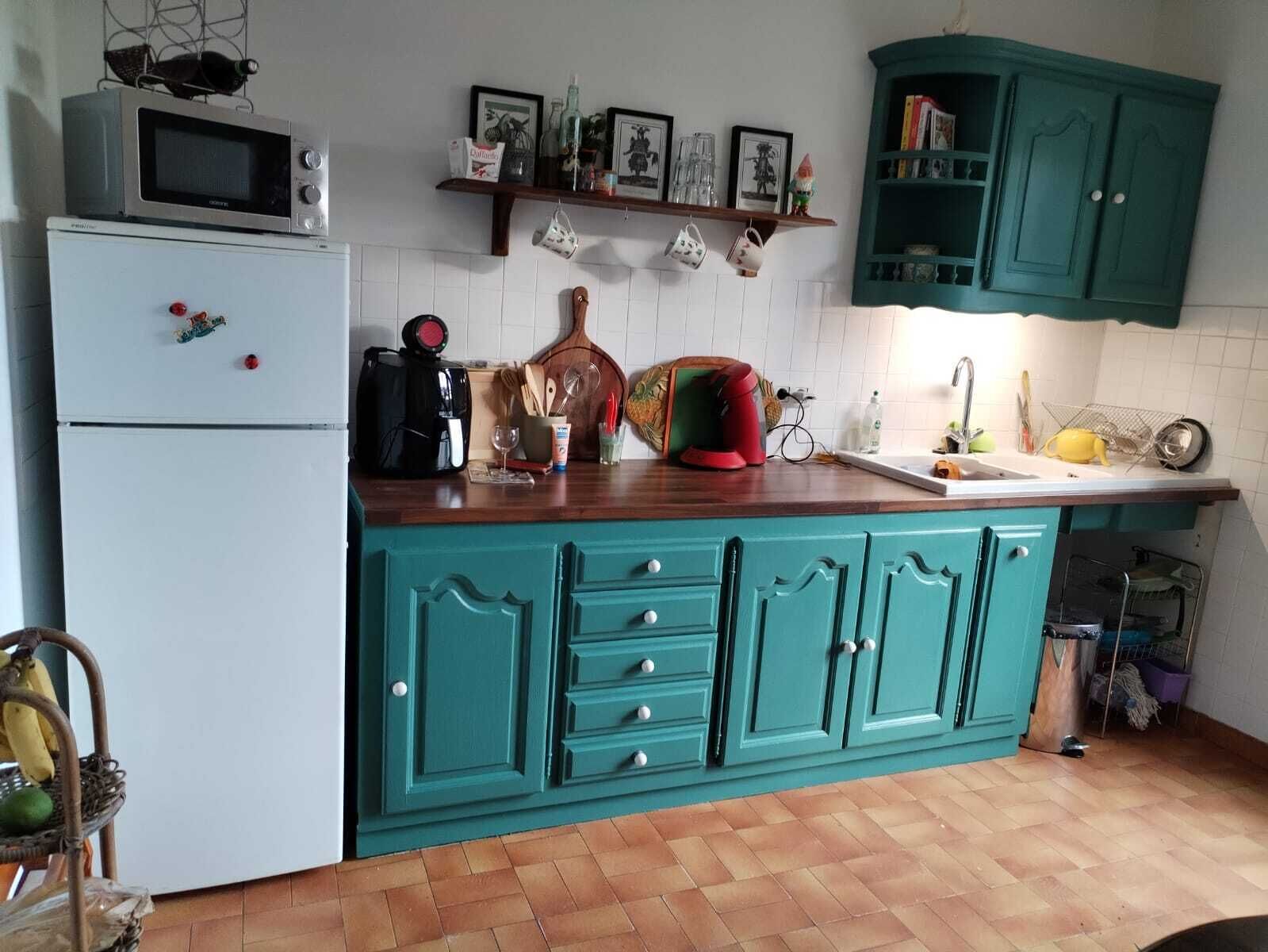 A kitchen with teal cabinets, a white refrigerator, a microwave, and a coffee maker. There are various kitchen utensils and a fruit basket on the counter.