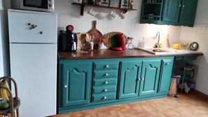 A kitchen with teal cabinets, a white refrigerator, a microwave, and a coffee maker. There are various kitchen utensils and a fruit basket on the counter.