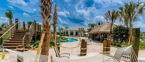 Uma área de piscina em resort com um edifício de telhado de palha, palmeiras e redes.
