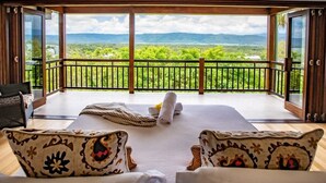A bedroom with a large bed, two patterned chairs, a balcony with a wooden railing, and a view of a green landscape. - Bangalow - Best View in Port Douglas (Port Douglas)