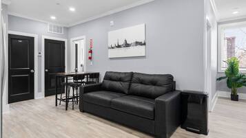 A modern living room with a black leather sofa, a dining table with chairs, and a black speaker.