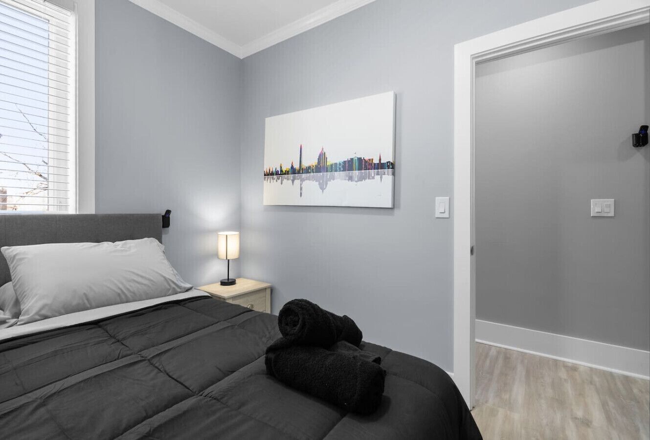 Une chambre à coucher avec un lit, une table de chevet, une lampe, une serviette enroulée et une œuvre d’art représentant un paysage urbain accrochée au mur.