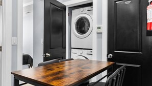 Une petite pièce avec une table et des chaises en bois, un lave-linge et une porte donnant sur le local du concierge.