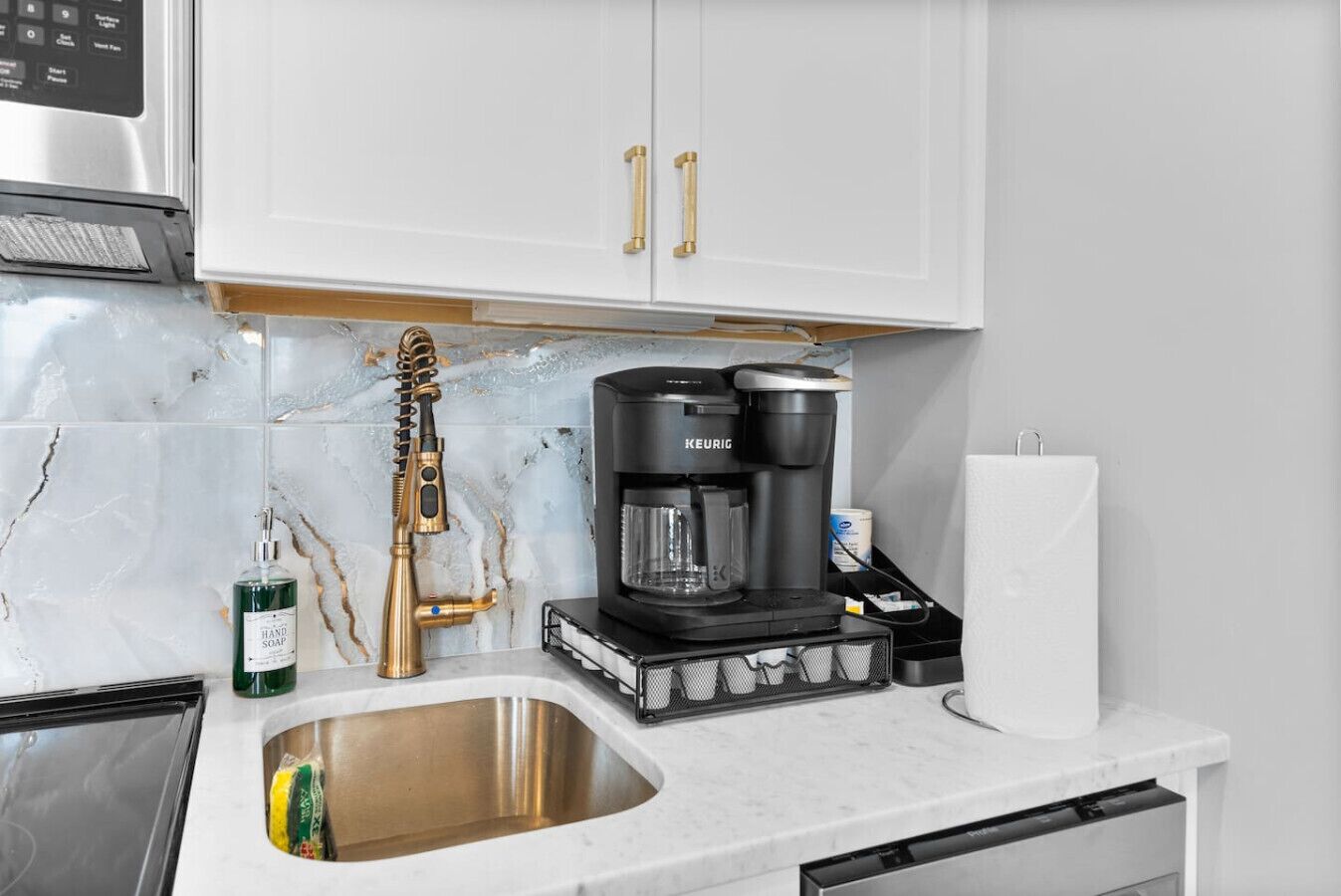 A kitchen with a stainless steel sink, a coffee maker, a hand soap dispenser, and a paper towel holder.