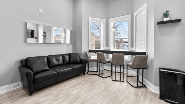 A modern living room with a black leather sofa, three grey chairs, a large mirror, and a window with blinds.