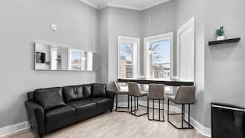 A modern living room with a black leather sofa, three grey chairs, a large mirror, and a window with blinds.