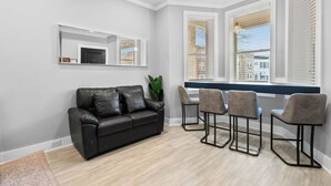 Un salon moderne avec un canapé en cuir noir, une table basse et quatre tabourets de bar. Il y a deux fenêtres équipées de stores, un luminaire au plafond et une petite étagère sur laquelle se trouve un téléviseur.