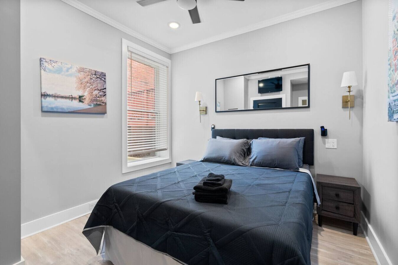 A bedroom with a bed, a window with blinds, a painting, a mirror, and a bedside table with a lamp.