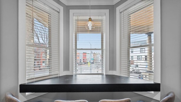 Un coin repas avec une table noire et quatre chaises, de grandes fenêtres équipées de stores et un luminaire suspendu.