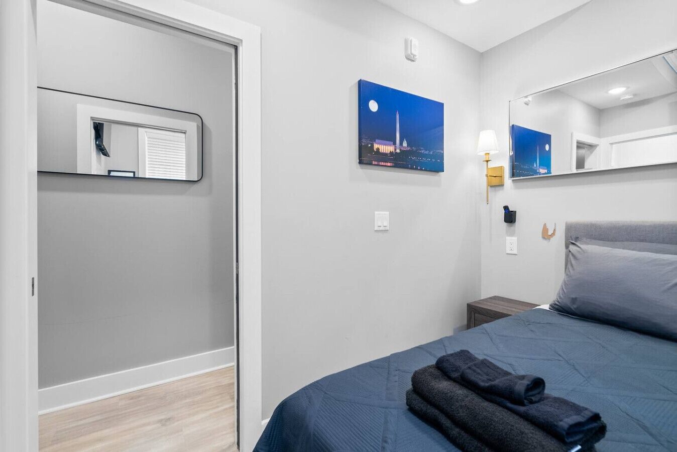 Habitación de hotel con cama, mesita de noche, lámpara, un cuadro de un paisaje urbano y una puerta con ventanal.