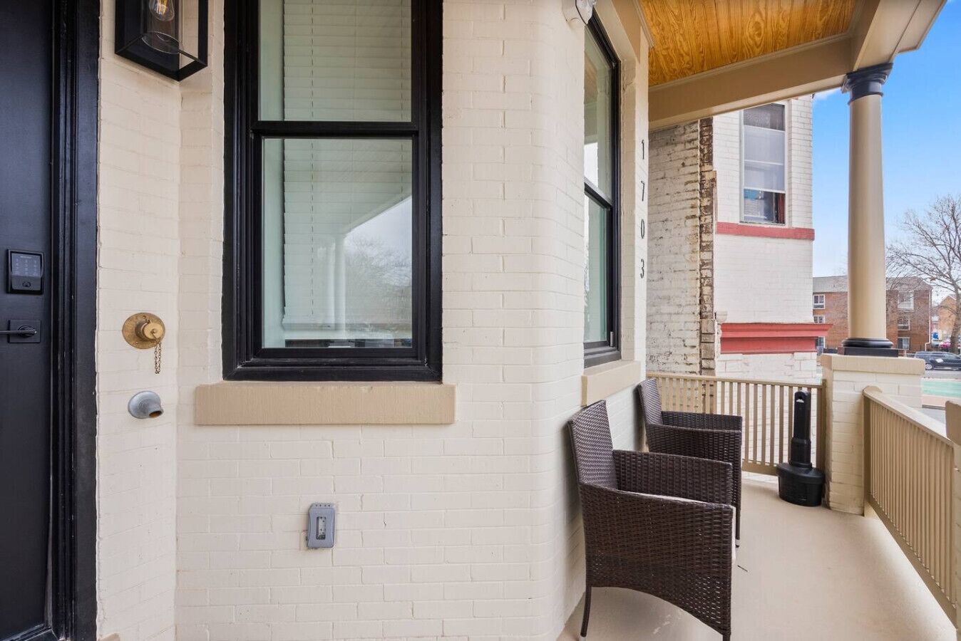 Fachada de edificio de ladrillo blanco, ventana con marco negro, puerta con cerradura y un pequeño área exterior con sillas de mimbre.