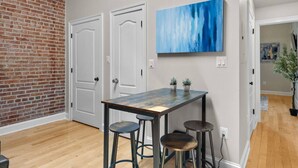 A room with a brick wall, two white doors, a blue abstract painting, a wooden table with stools, and a potted plant.