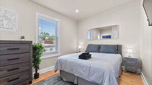 A bedroom with a bed, dresser, bedside tables, a potted plant, and a window with blinds.