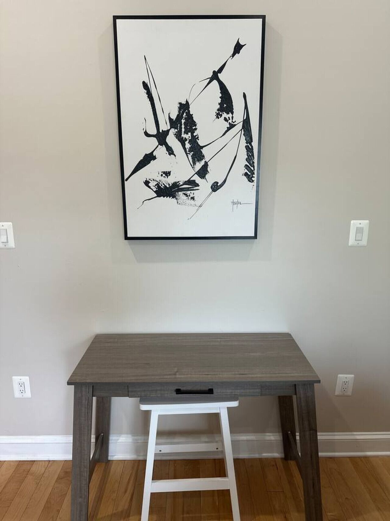 Un tableau abstrait encadré est accroché au mur, au-dessus d’un bureau en bois, devant lequel se trouve un tabouret blanc.
