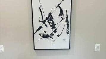 Un tableau abstrait encadré est accroché au mur, au-dessus d’un bureau en bois, devant lequel se trouve un tabouret blanc.