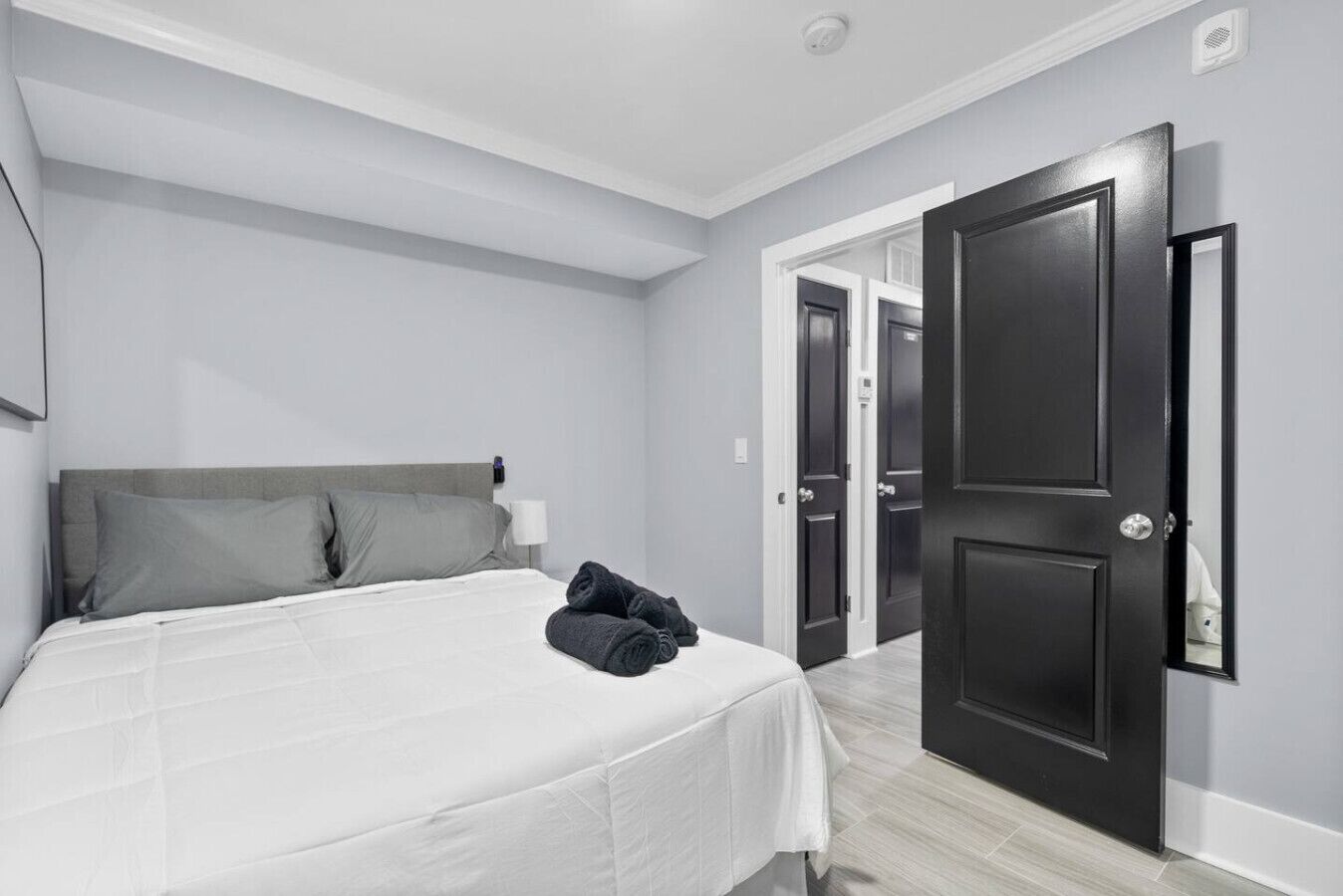 Un dormitorio con cama, almohadas grises, una colcha blanca y una toalla enrollada sobre la cama. Hay dos puertas, una abierta y otra cerrada, que dan a otra habitación.