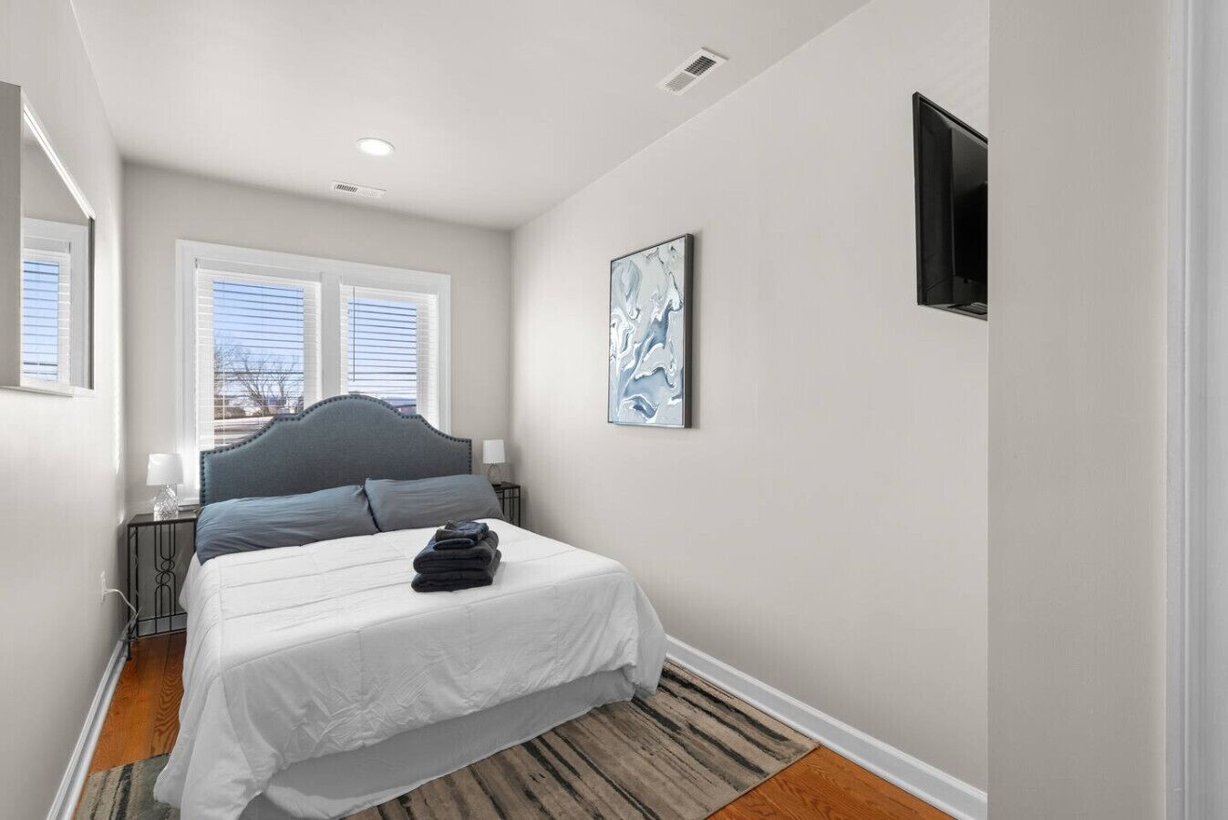 Une chambre à coucher comprenant un lit, deux tables de chevet, une fenêtre avec des stores, un luminaire au plafond, un téléviseur fixé au mur et un tableau encadré.