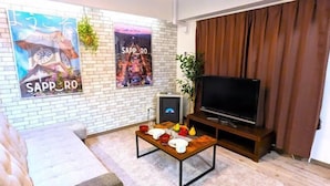 A living room with a brick wall, a sofa, a wooden coffee table with dishes, a TV on a wooden stand, and a poster on the wall. - KIYAZA Station Sapporo 2 (Sapporo)