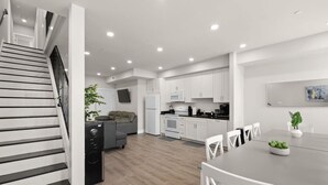 Eine moderne Küche mit weißen Schränken, ein Essbereich mit einem grauen Tisch und weißen Stühlen sowie eine Treppe mit schwarzen Stufen.
