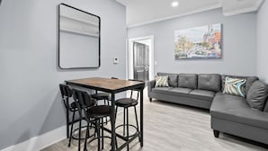 Un salon moderne avec un canapé gris, une table à manger en bois et des chaises, ainsi qu’un tableau représentant un paysage urbain accroché au mur.