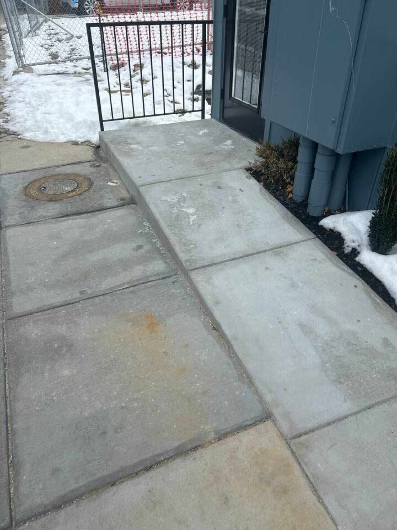 Ein Betonweg führt zu einem Gebäudeeingang mit einem Metallgitter, einem kleinen Baum und einem mit Schnee bedeckten Boden.