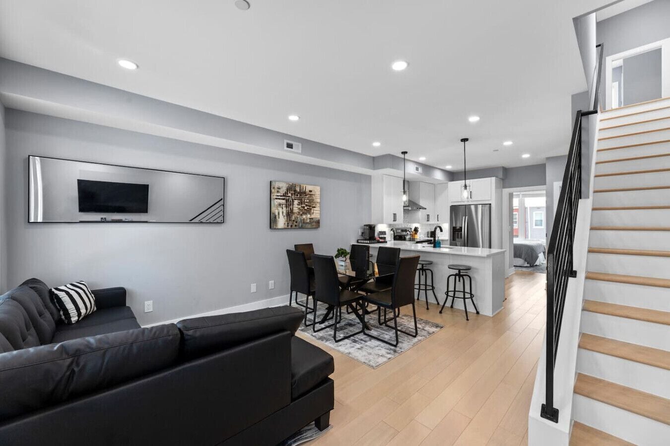 A modern living room with a black sofa, a dining area with a table and chairs, a kitchen with a stainless steel refrigerator, and a staircase with wooden steps.