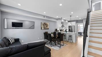 A modern living room with a black sofa, a dining area with a table and chairs, a kitchen with a stainless steel refrigerator, and a staircase with wooden steps.