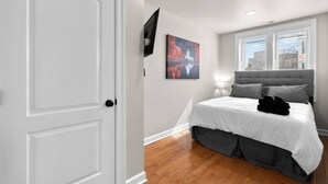 Un dormitorio con una cama, un televisor en la pared, una ventana con persianas y una puerta.
